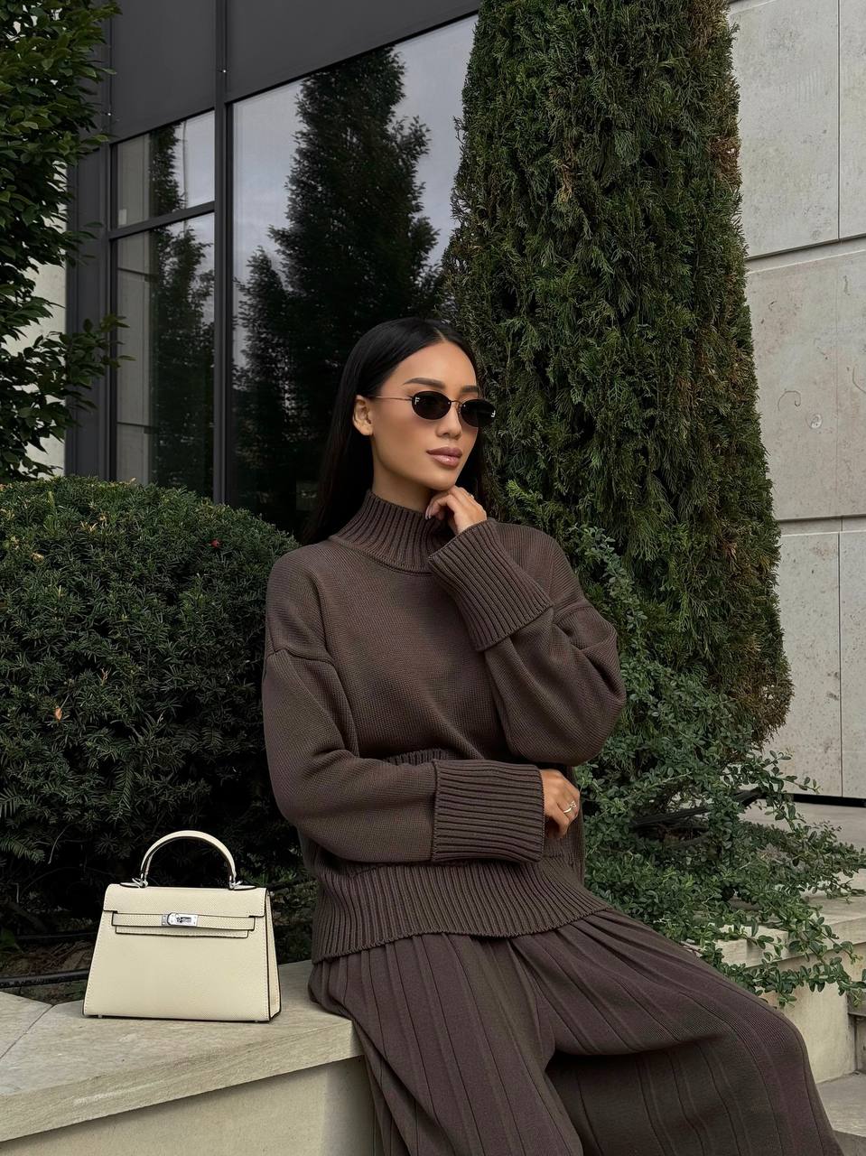 Woman in a brown outfit with a beige handbag standing outdoors.