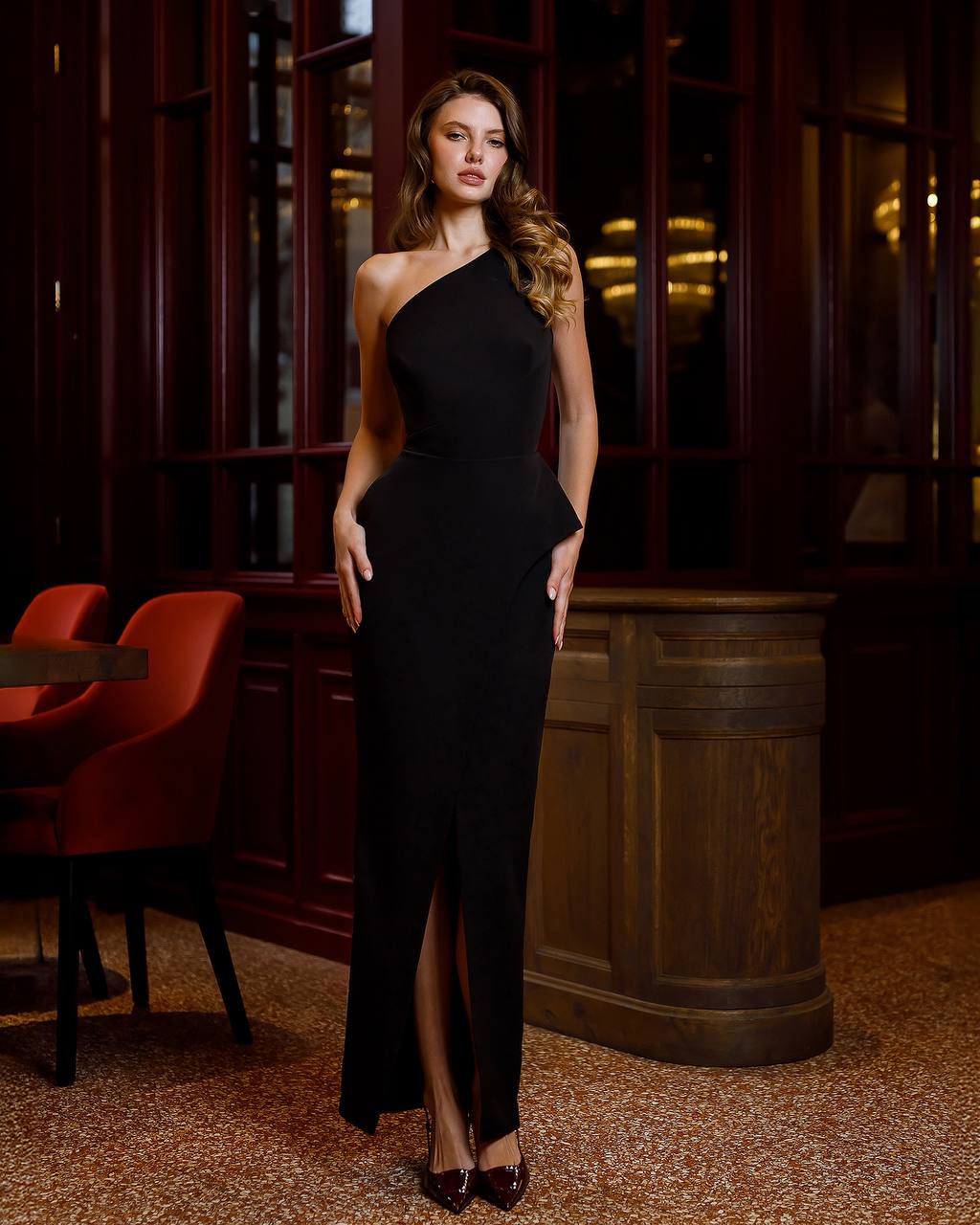 Woman in a black one-shoulder dress standing in an elegant interior setting.