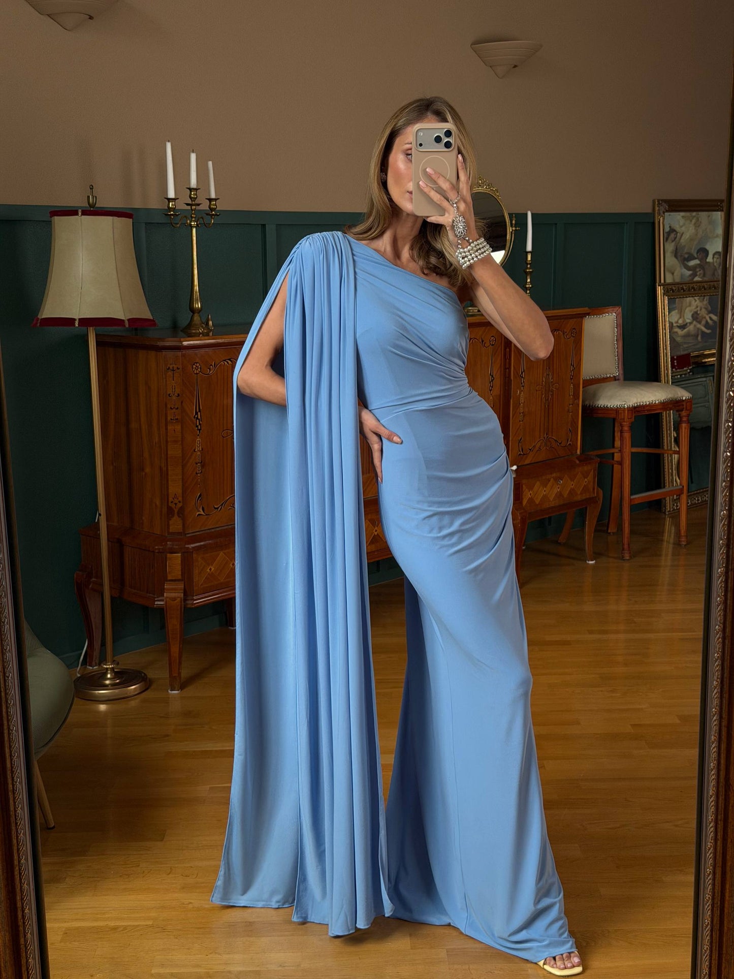 Woman in a blue evening gown standing in a room with wooden furniture and a lamp.