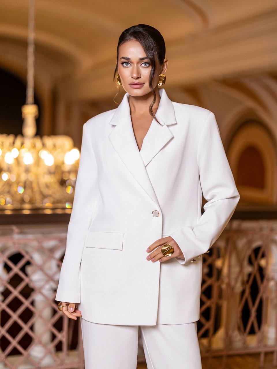 Woman in a white suit standing in an elegant interior setting with chandeliers.