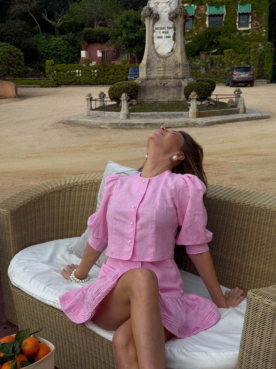Woman in a pink dress sitting on a wicker chair outdoors with a statue in the background.