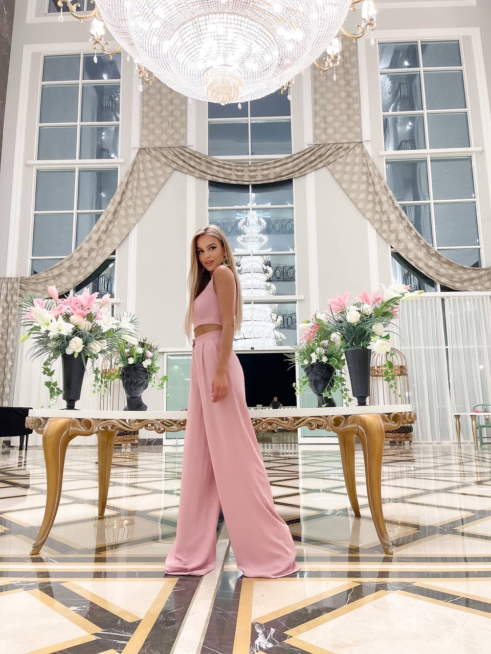 Woman in a pink outfit standing in a luxurious interior setting with chandelier and floral arrangements.