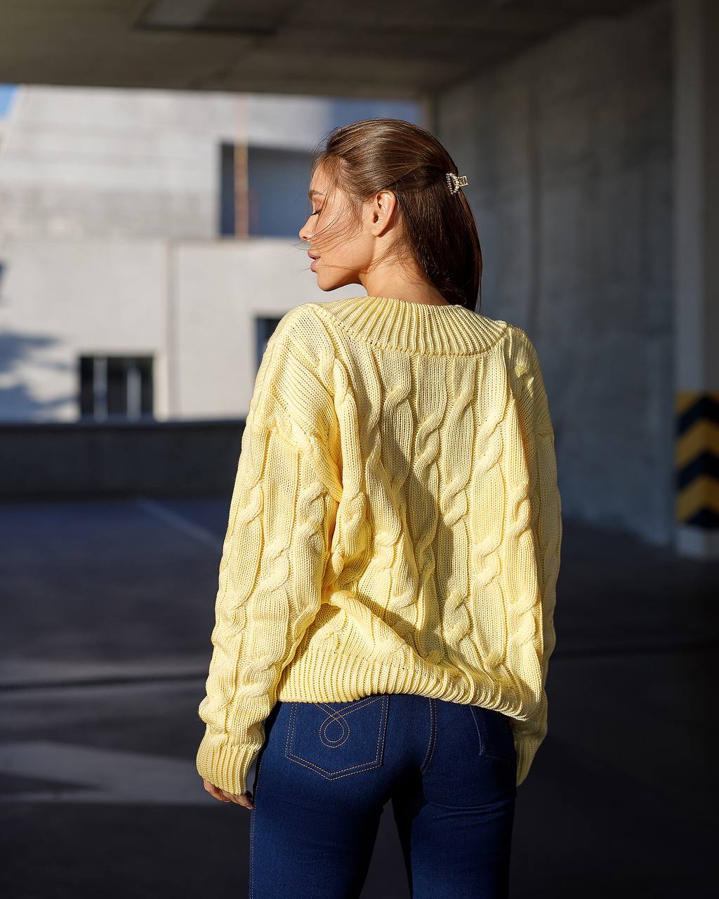 Woman wearing a yellow cable knit sweater and blue jeans standing in an outdoor setting.