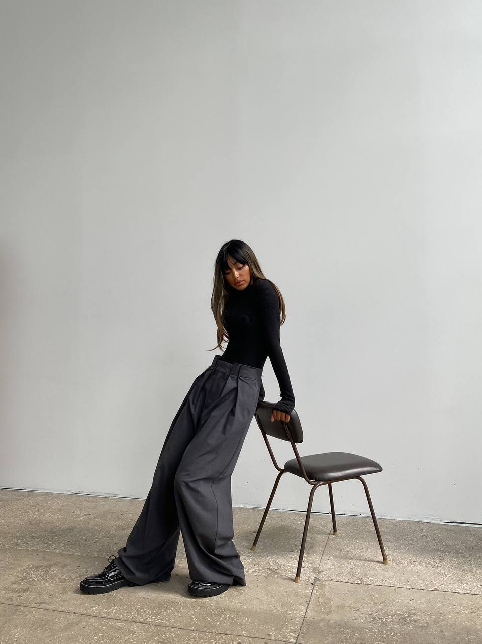 Person wearing a black top and gray wide-leg pants standing next to a chair against a plain wall.