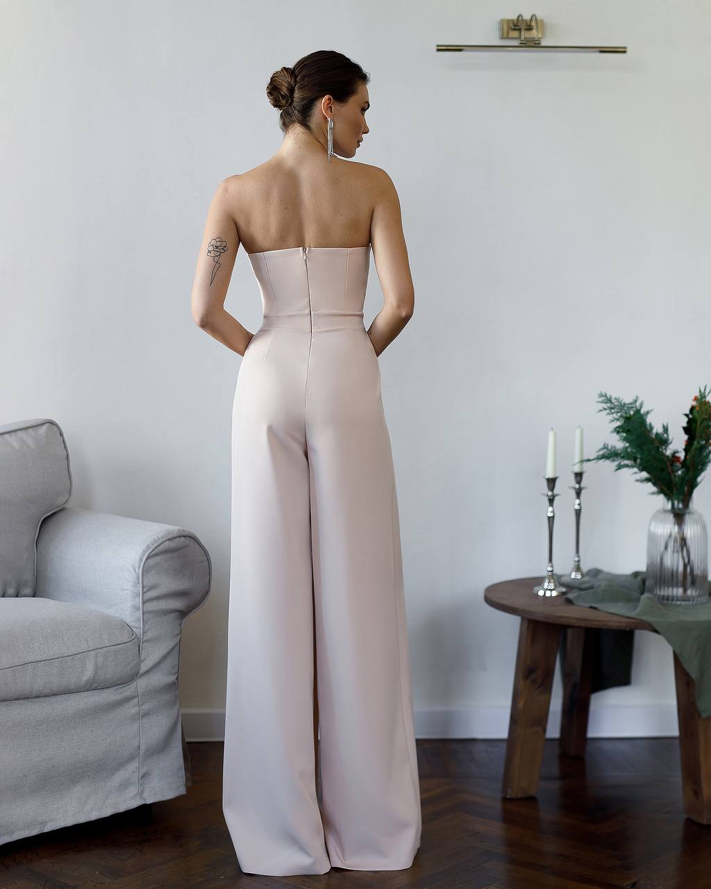 Woman wearing a strapless beige jumpsuit in a room with a gray armchair and wooden table.