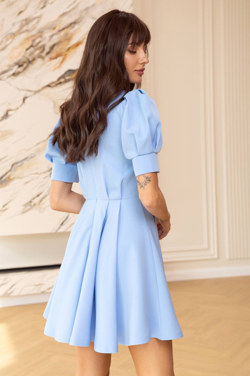 Woman wearing a light blue dress standing in a room with a stone wall and wooden floor.