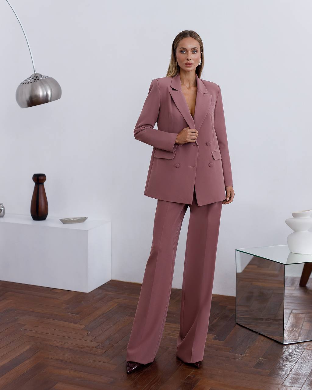 Woman in a pink suit standing in a modern room with wooden flooring and white walls.