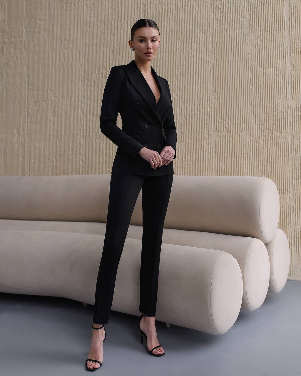 Woman in a black suit standing in front of a modern beige sofa against a textured wall.