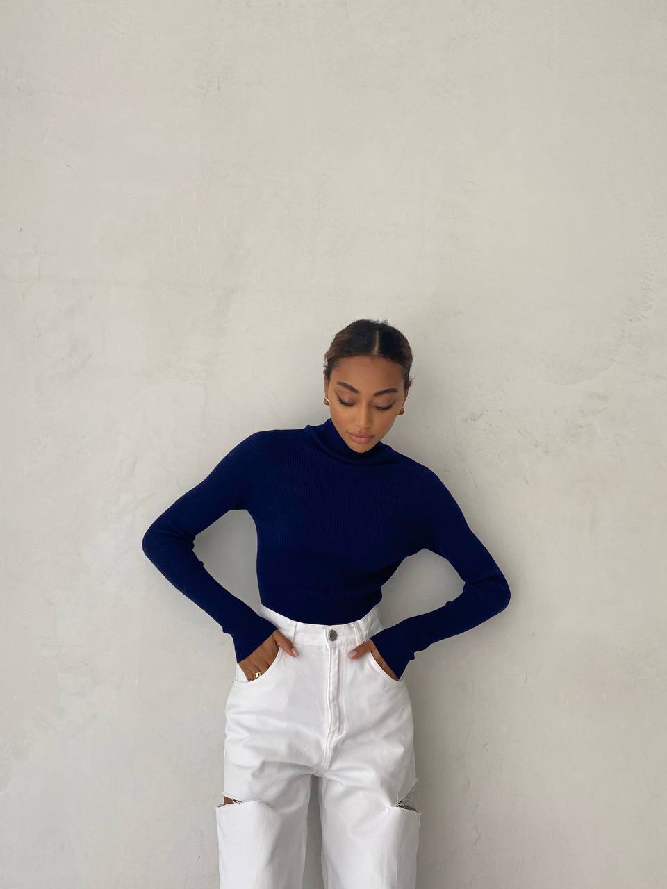 Person wearing a navy blue turtleneck and white pants against a plain background