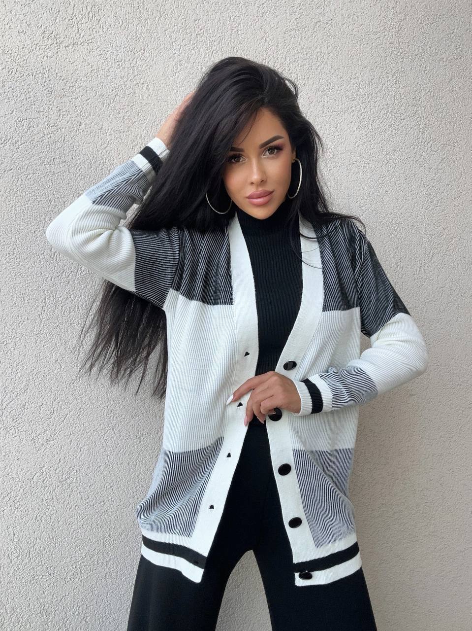 Woman wearing a white and gray cardigan over a black outfit against a plain background