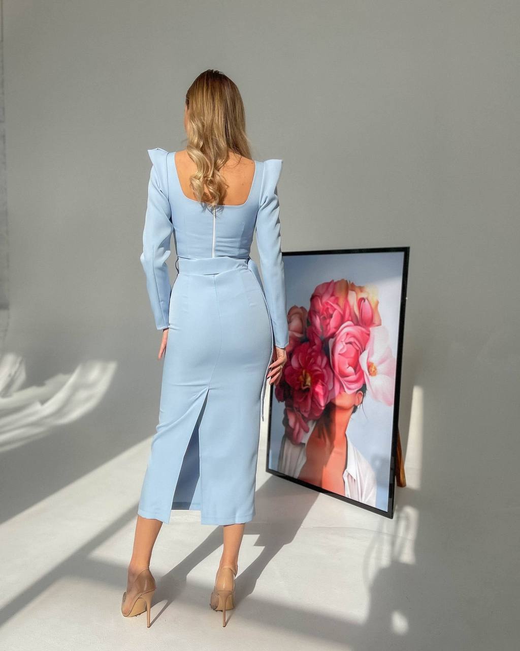 Woman in a light blue dress standing in a sunlit room with a framed picture of flowers on the wall.