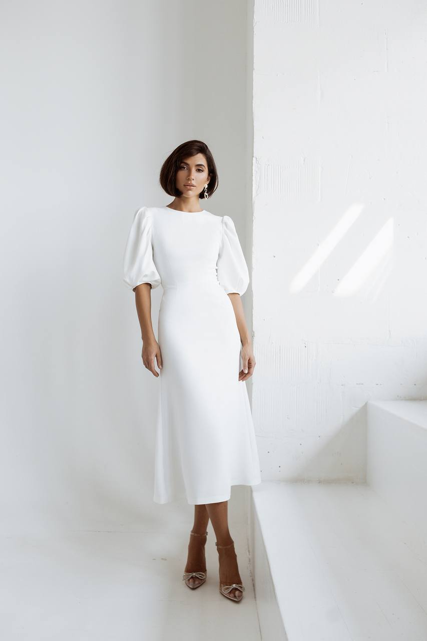 Woman wearing a white dress standing in a minimalistic setting with white walls and steps.