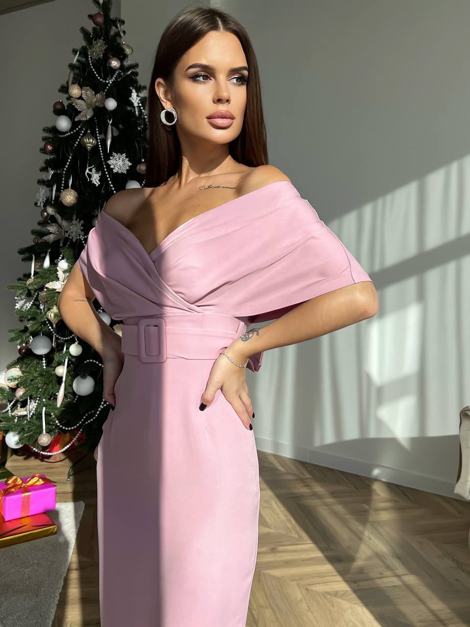 Woman in a pink off-shoulder dress standing in a room with a decorated Christmas tree.