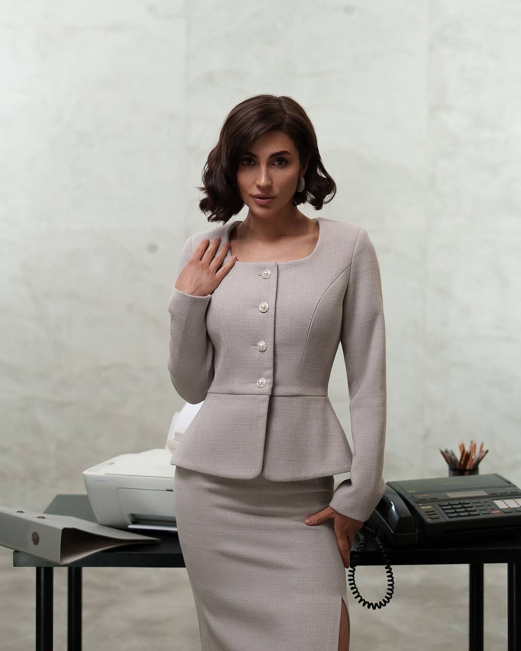 Woman in a light gray suit standing in an office setting with a printer and phone on a desk.