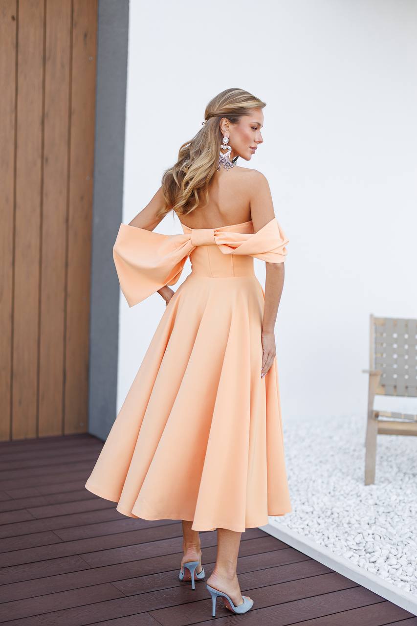 Woman in a peach dress standing on a wooden deck with a white wall and chair in the background.