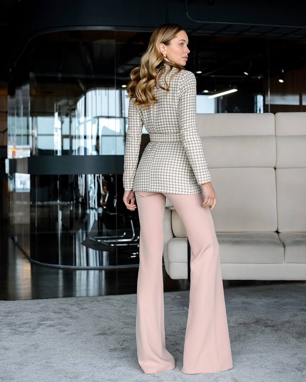 Woman in a checkered blazer and pink pants standing in a modern indoor setting.