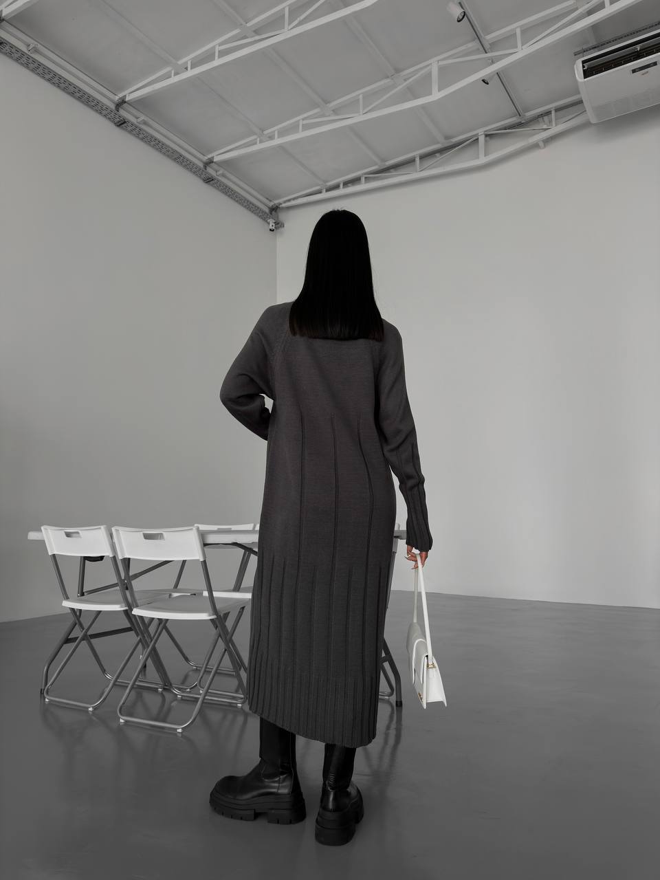 Person wearing a long dark coat standing in a minimalistic room with gray walls and floor.