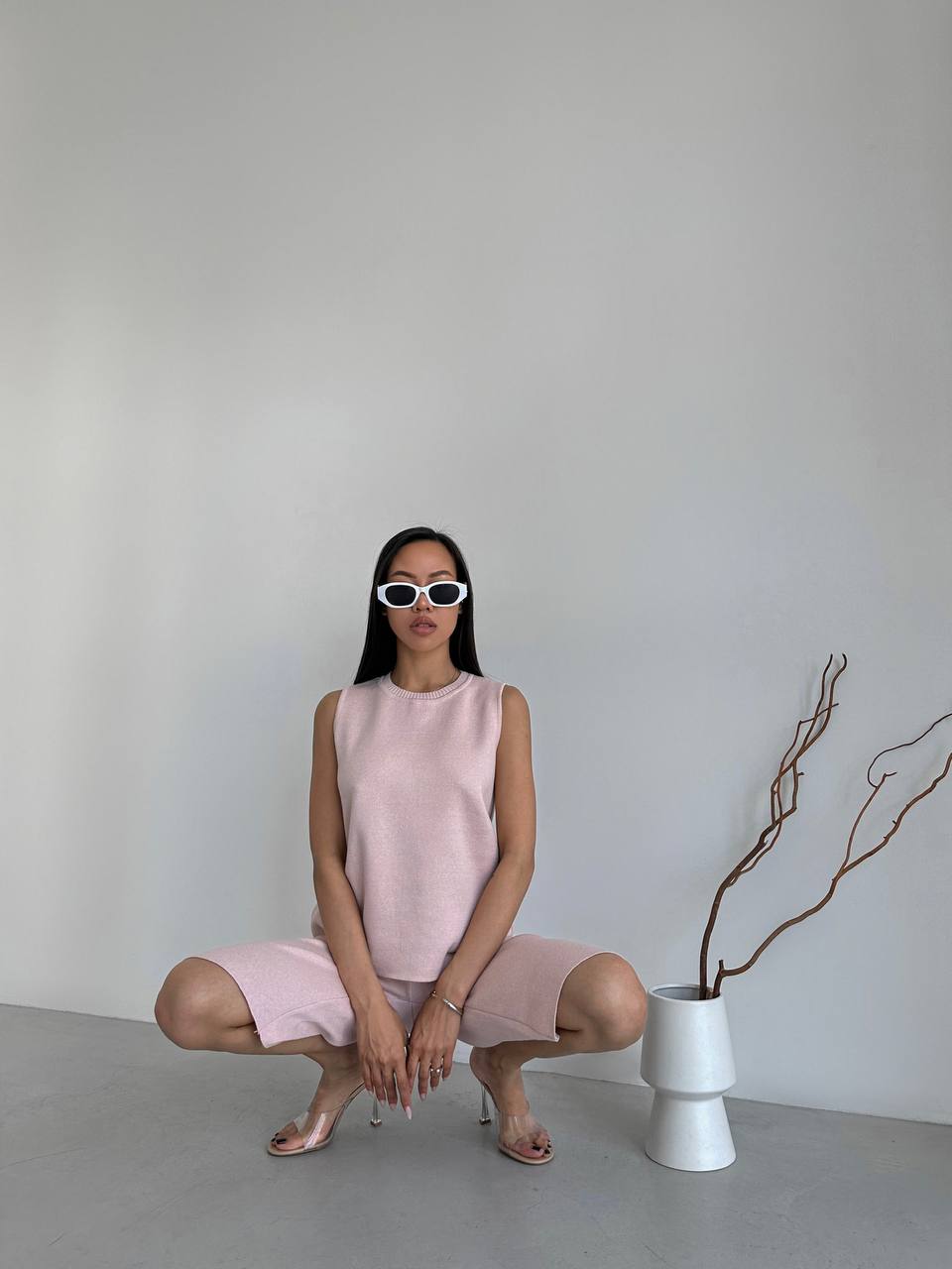 Woman in a pink dress sitting on the floor next to a white vase with a plant against a plain wall.