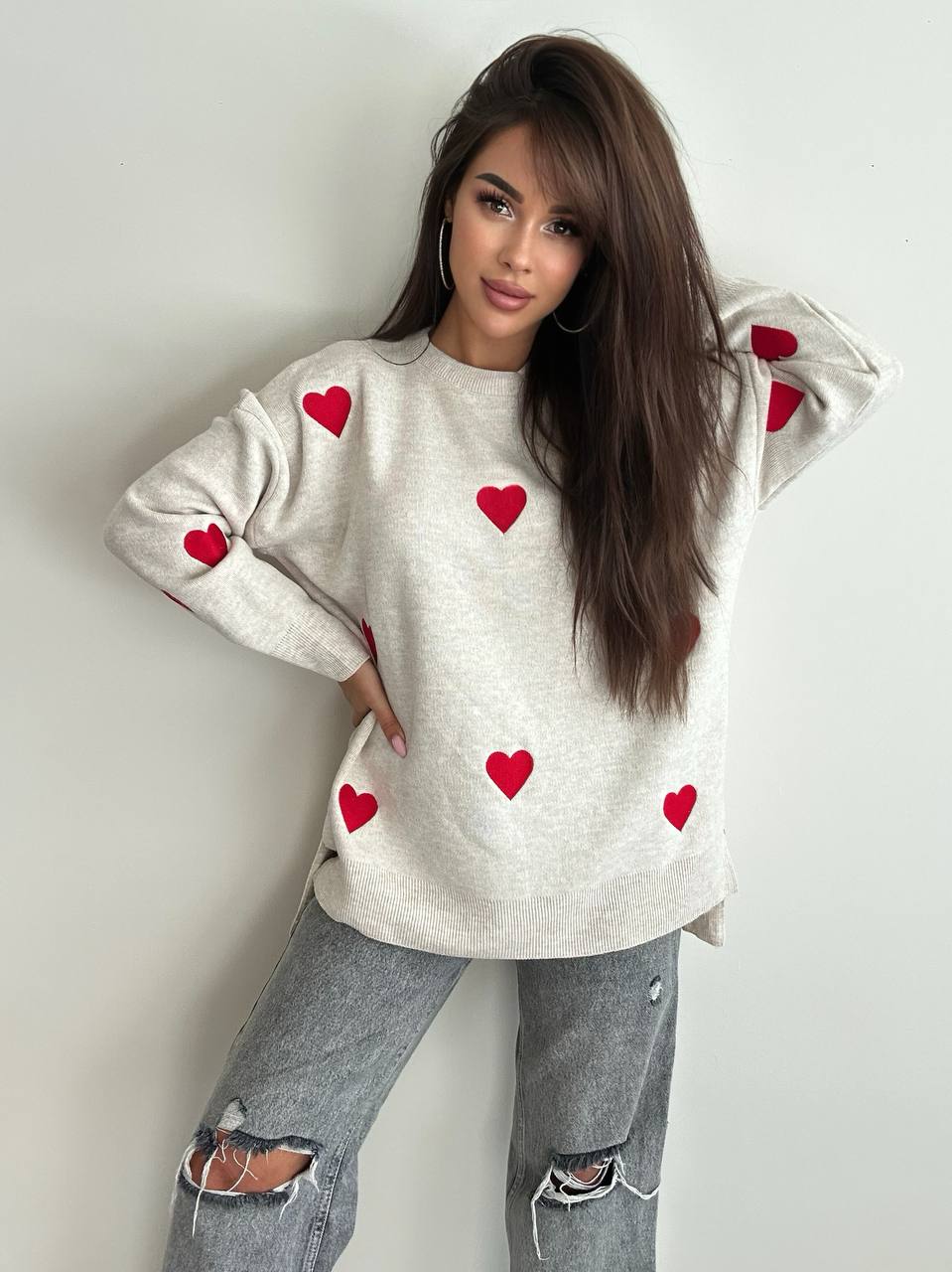Woman wearing a beige sweater with red heart patterns and ripped jeans against a plain background