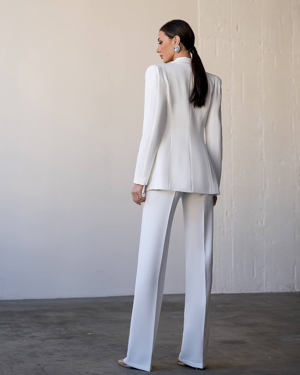 Woman wearing a white suit standing against a plain wall.
