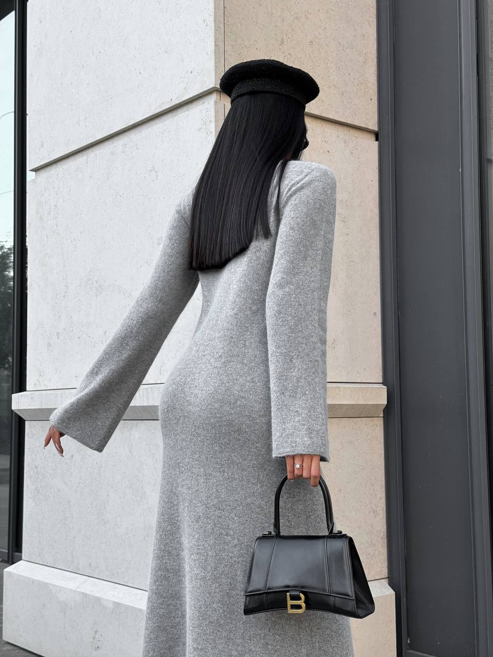Person in a gray outfit holding a black handbag, standing against a light-colored wall.