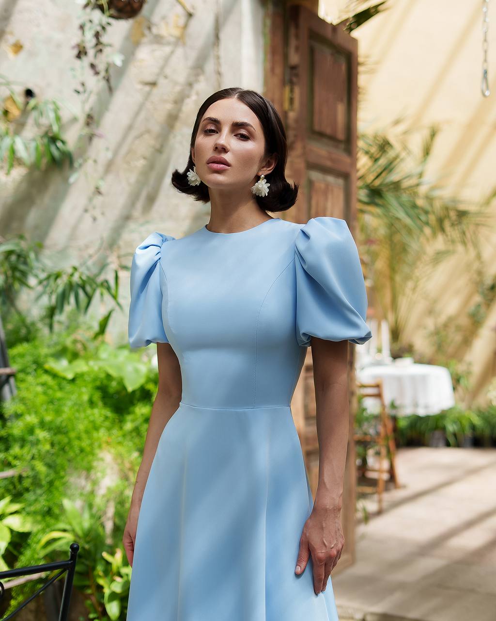 Woman in a light blue dress standing outdoors with greenery and a wooden structure in the background