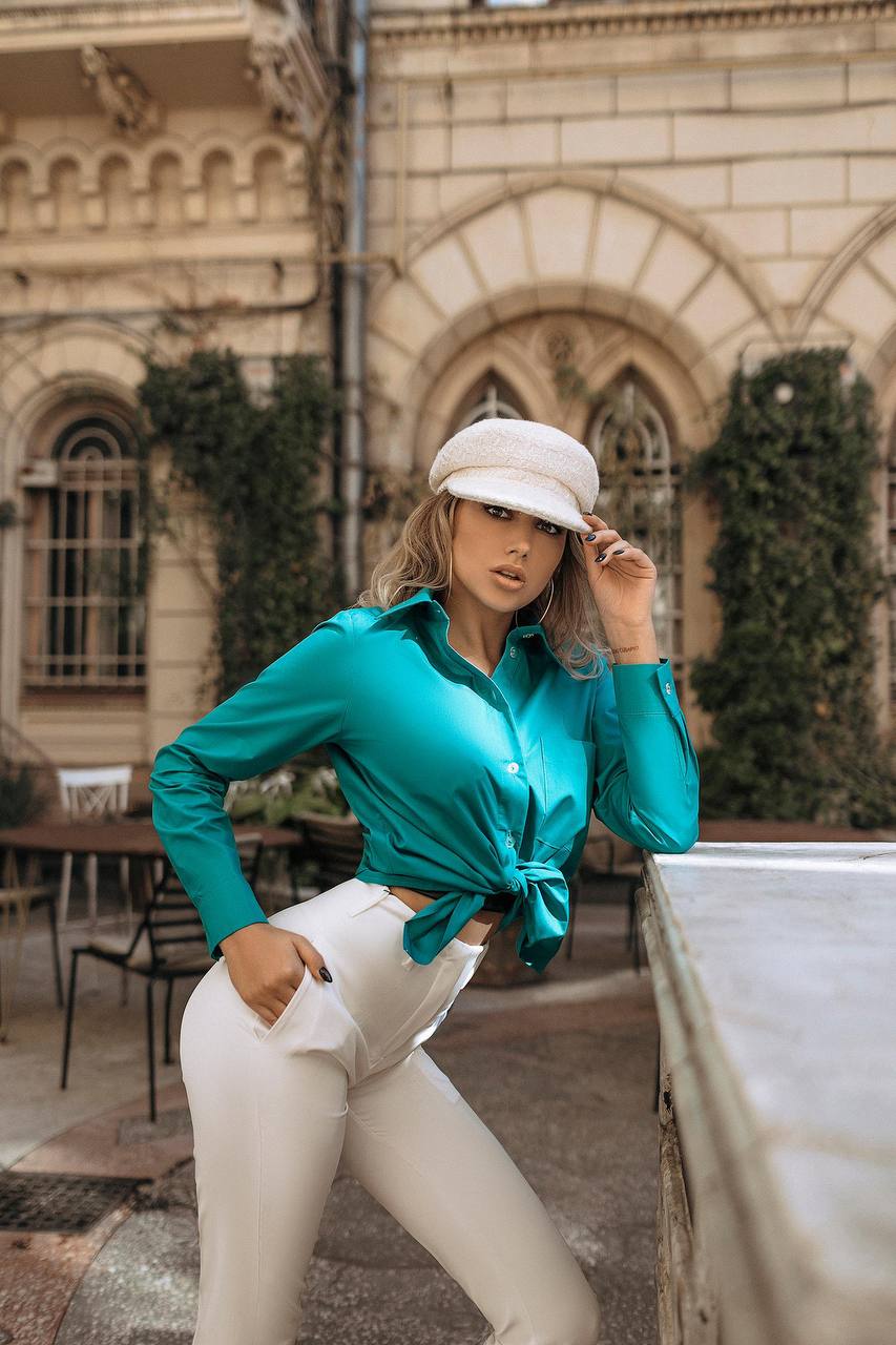 Woman in teal shirt and white pants standing outdoors with a building in the background