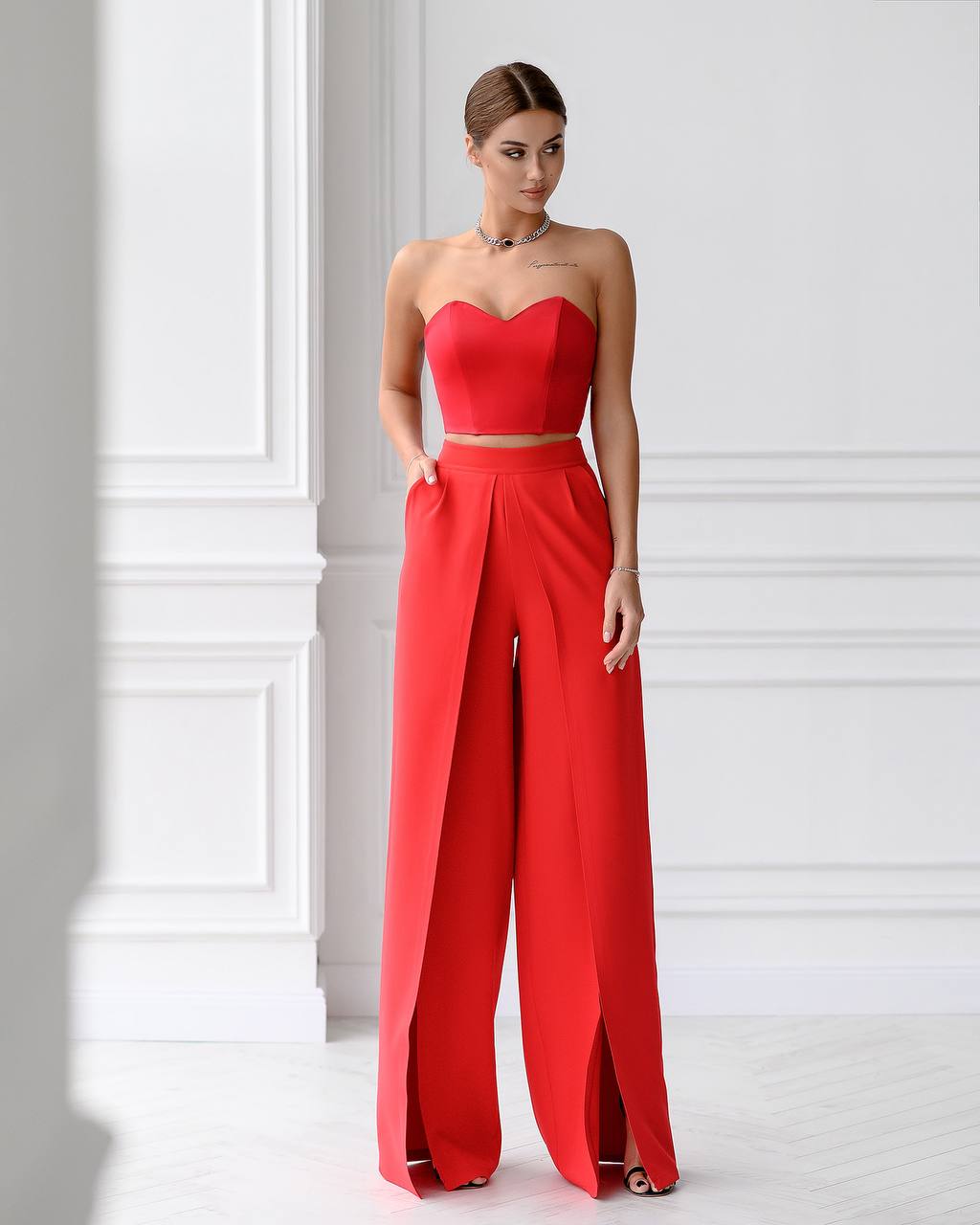 Woman wearing a red strapless top and wide-leg pants against a white wall.