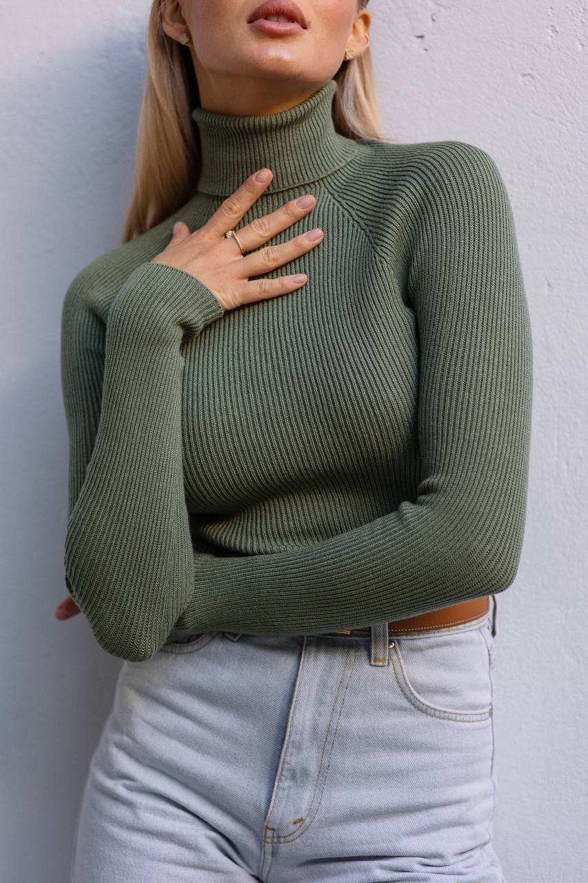 Person wearing a green ribbed sweater against a white wall