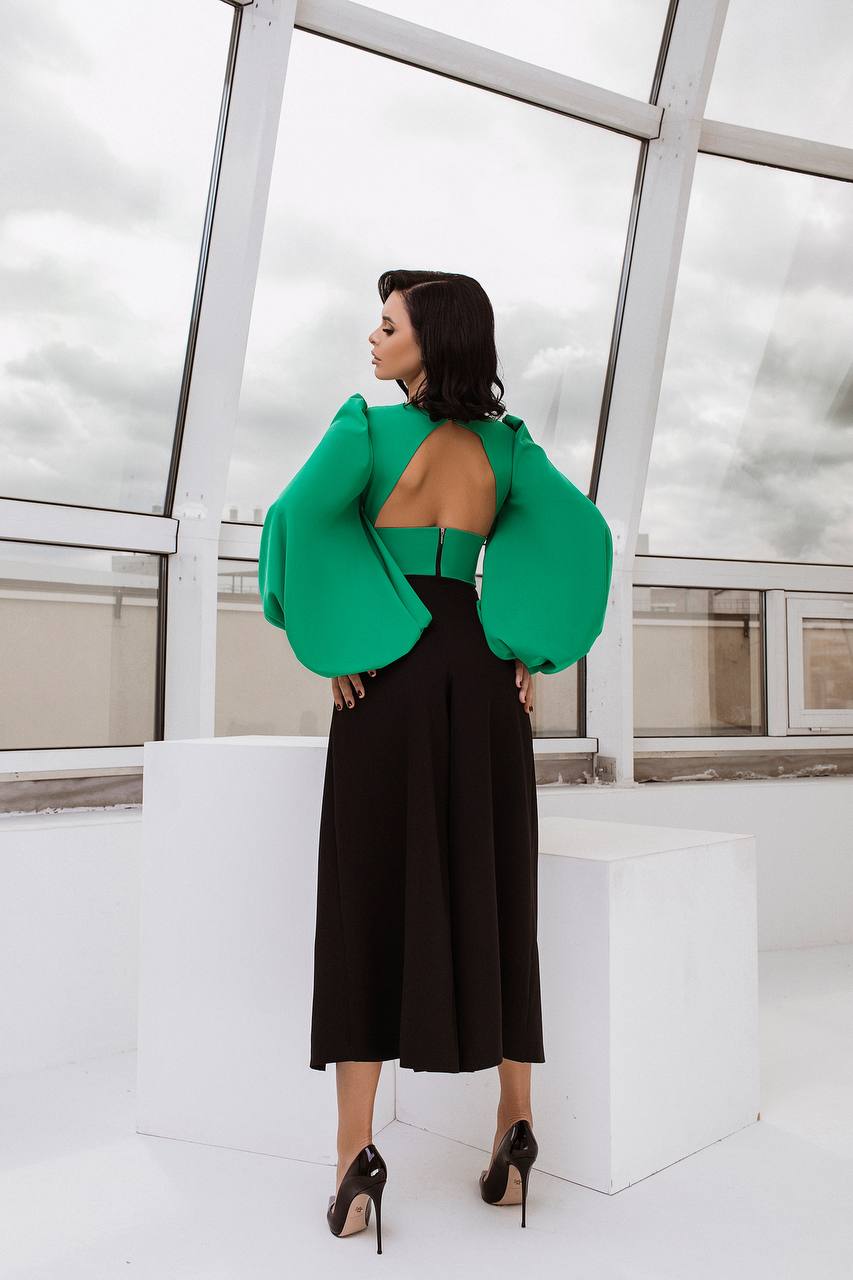 Woman wearing a green open-back top and black pants in an indoor setting with large windows.
