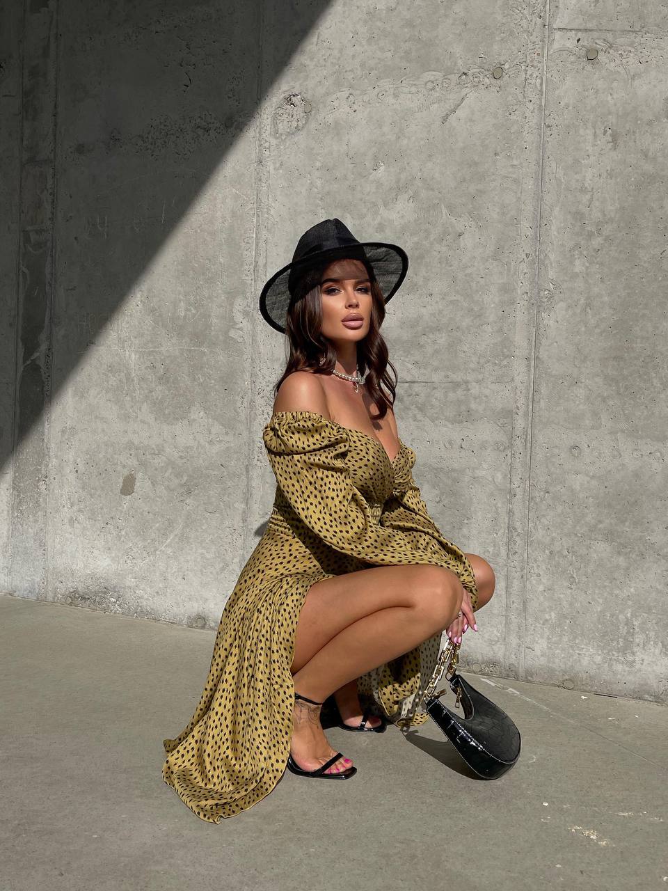 Woman in a yellow patterned dress and black hat posing against a concrete wall.
