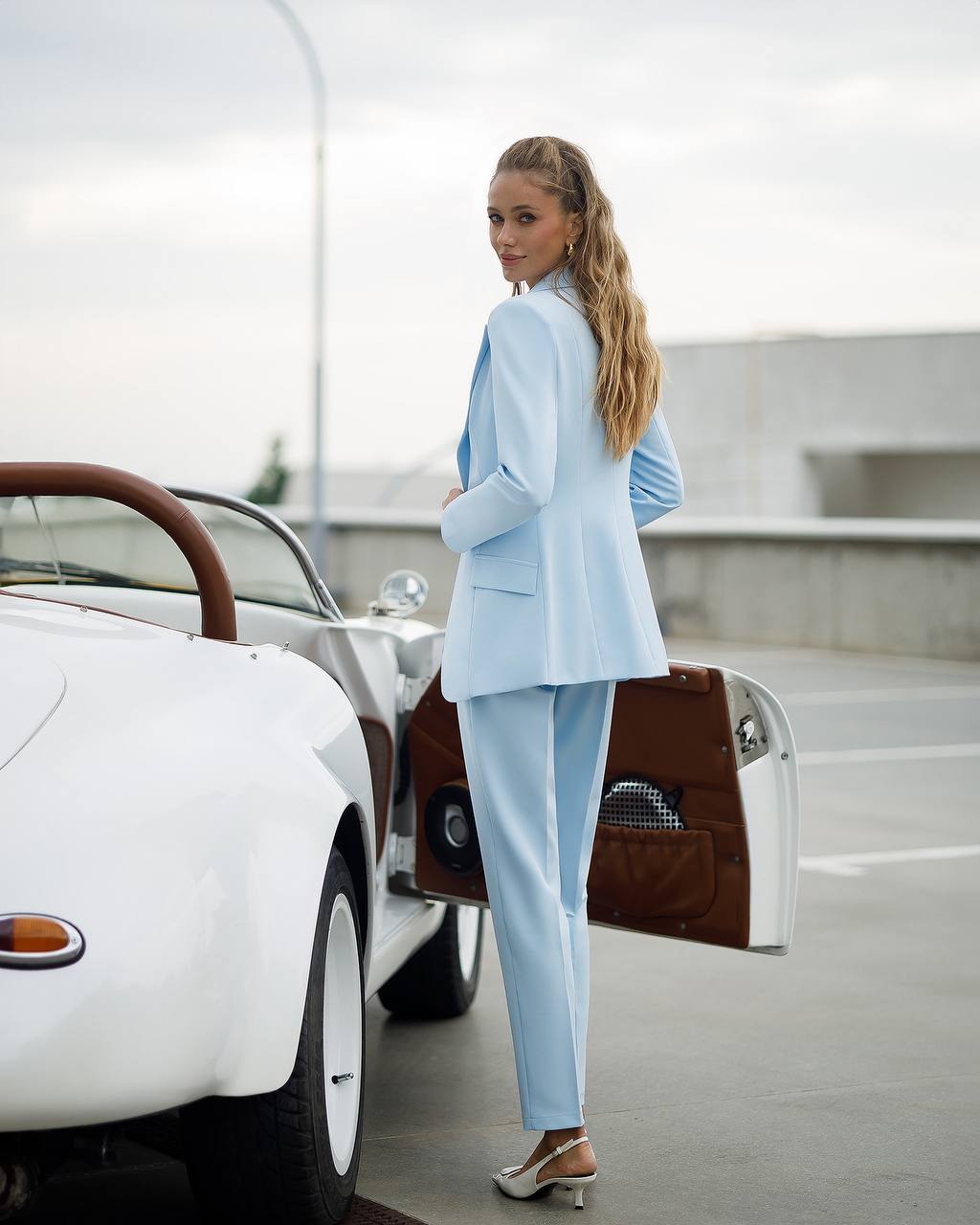 Woman in a light blue suit standing next to a vintage car in an indoor setting