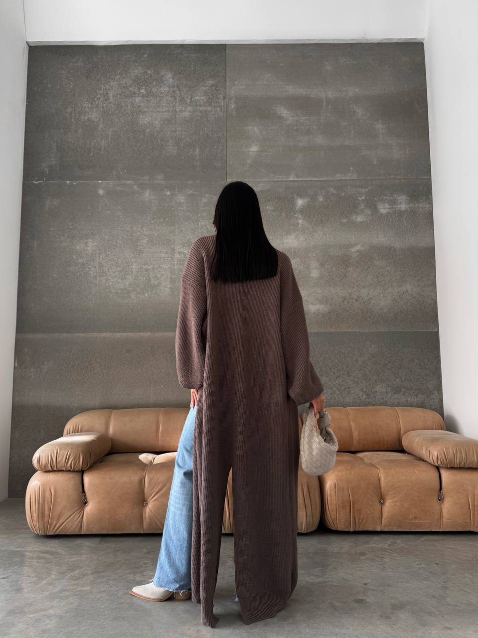 Person wearing a long brown coat standing in a room with a large abstract painting and brown sofa.