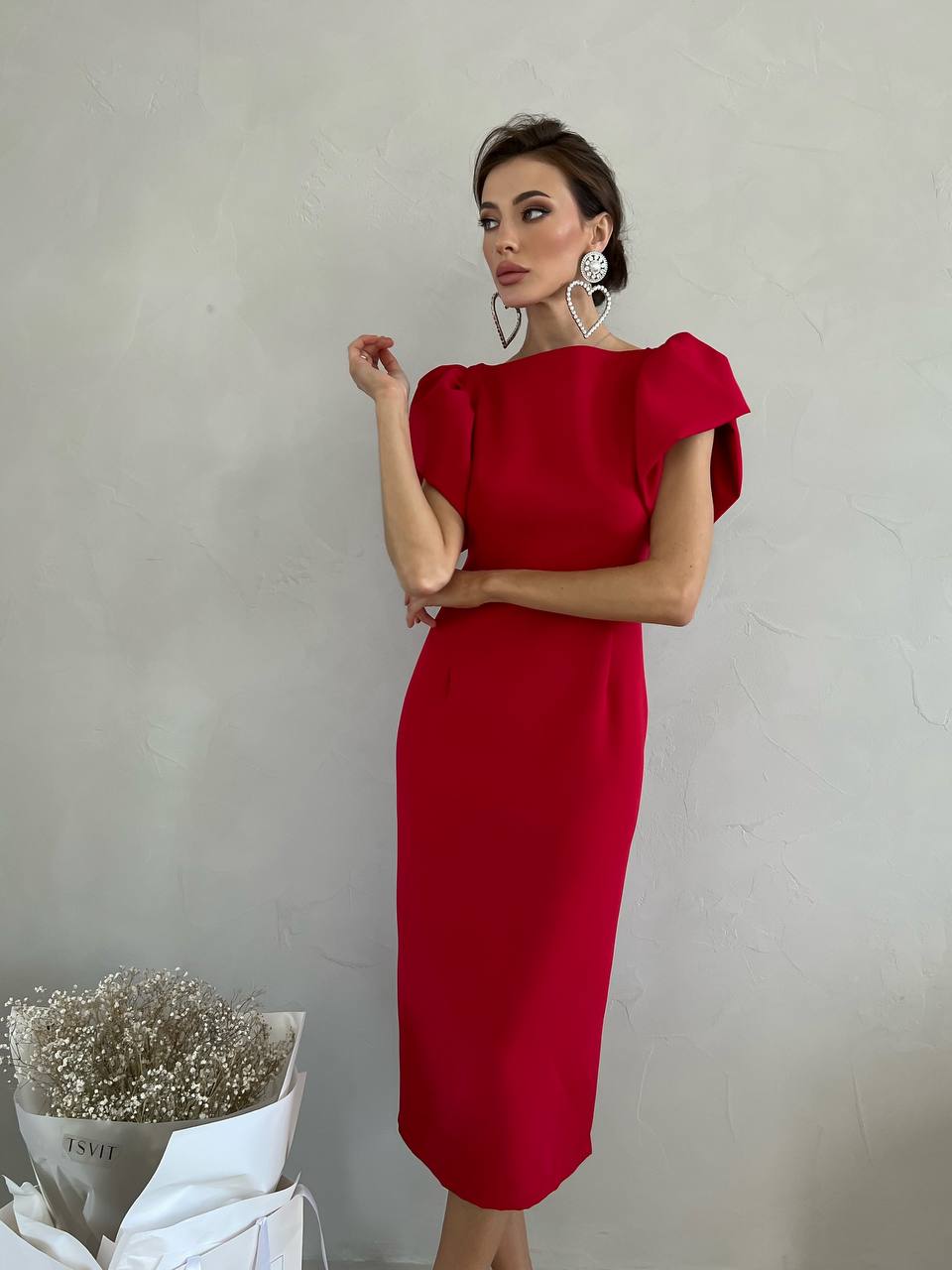 Woman wearing a red dress standing against a plain background with a vase of flowers on the left.