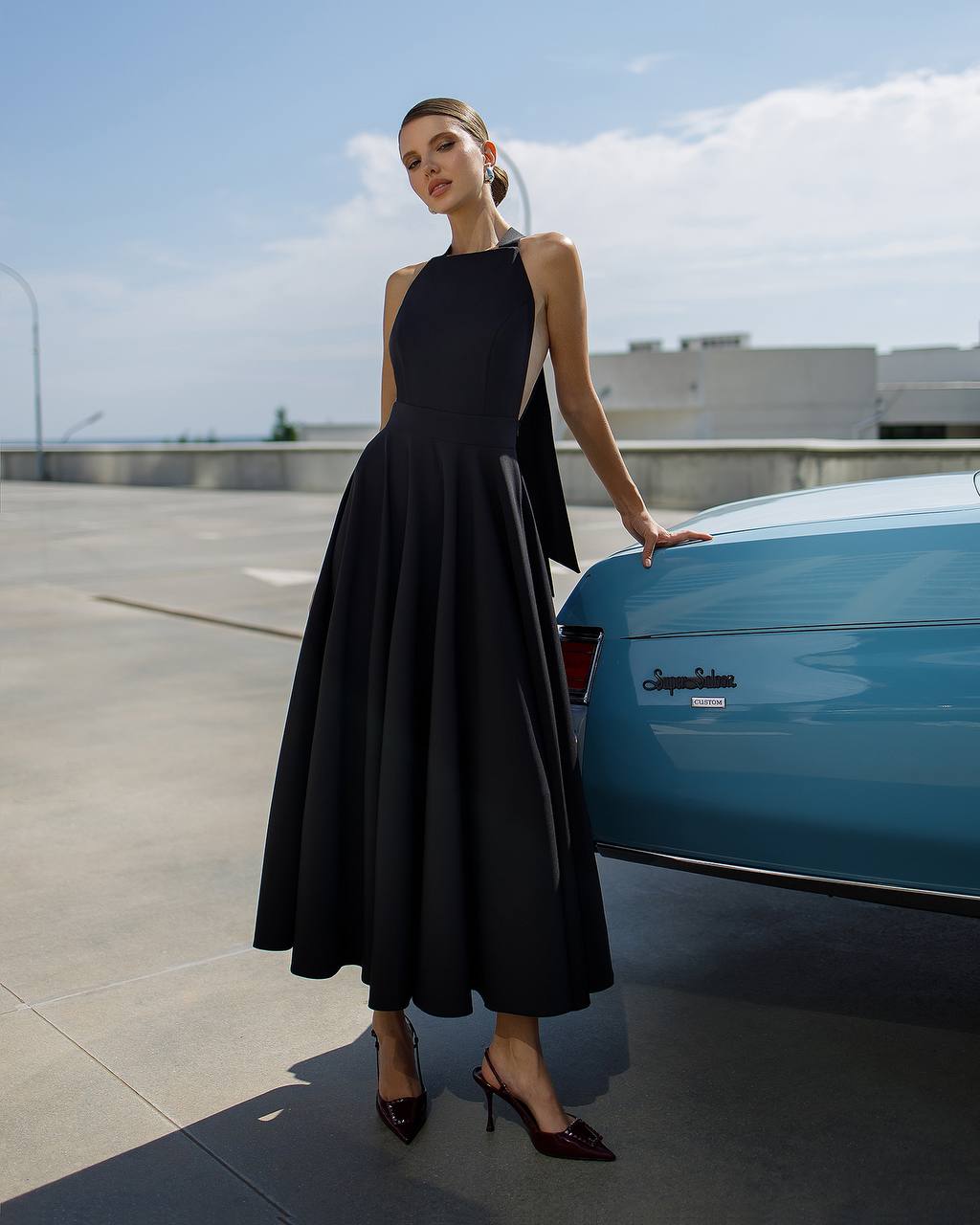 Woman in a black dress standing next to a blue car on a rooftop.