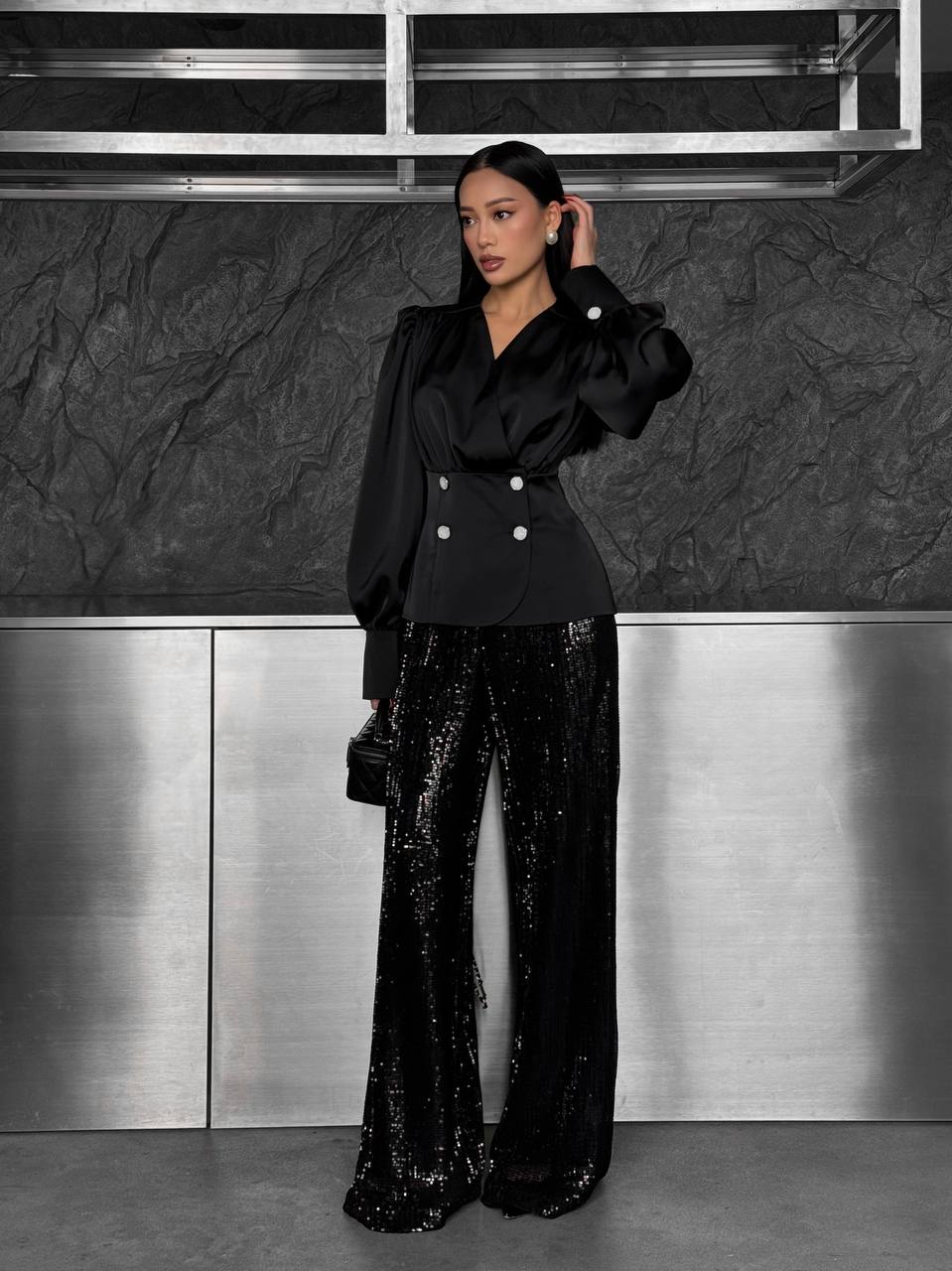 Woman in a black outfit with sequin pants standing against a metallic wall.