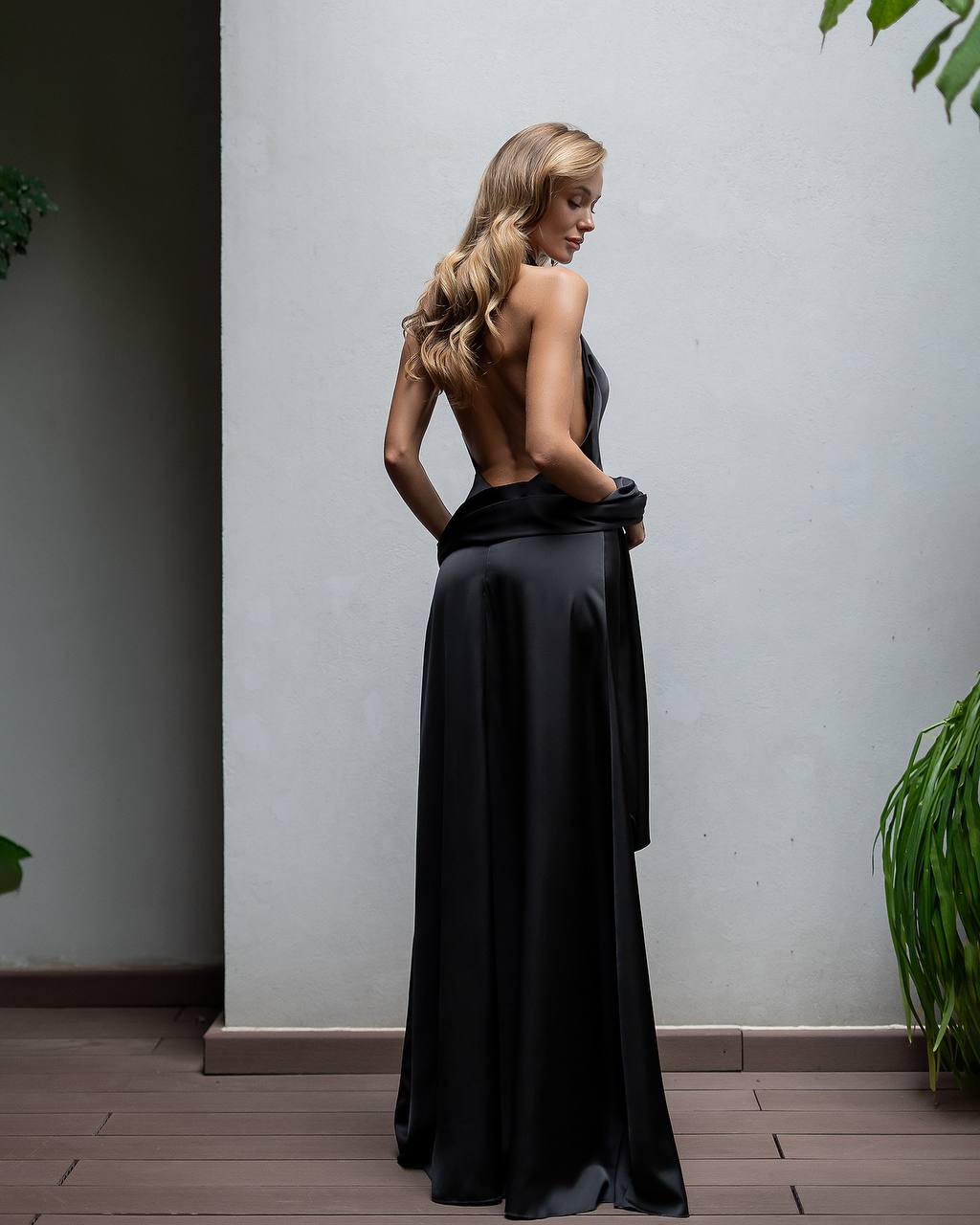 Woman in a black evening gown standing against a plain wall with plants around.