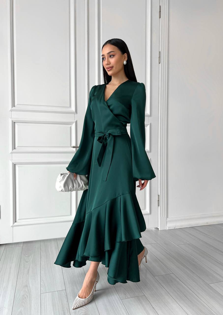 Woman wearing a green dress with long sleeves and ruffled hem in a room with white walls and wooden floor.