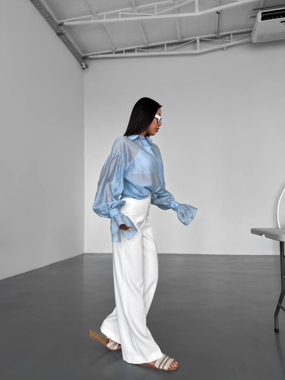 Person wearing a light blue sheer blouse and white pants in a minimalistic room.
