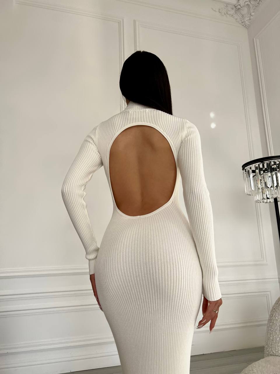Person wearing a white ribbed dress with an open back in a room with white walls and a chandelier.