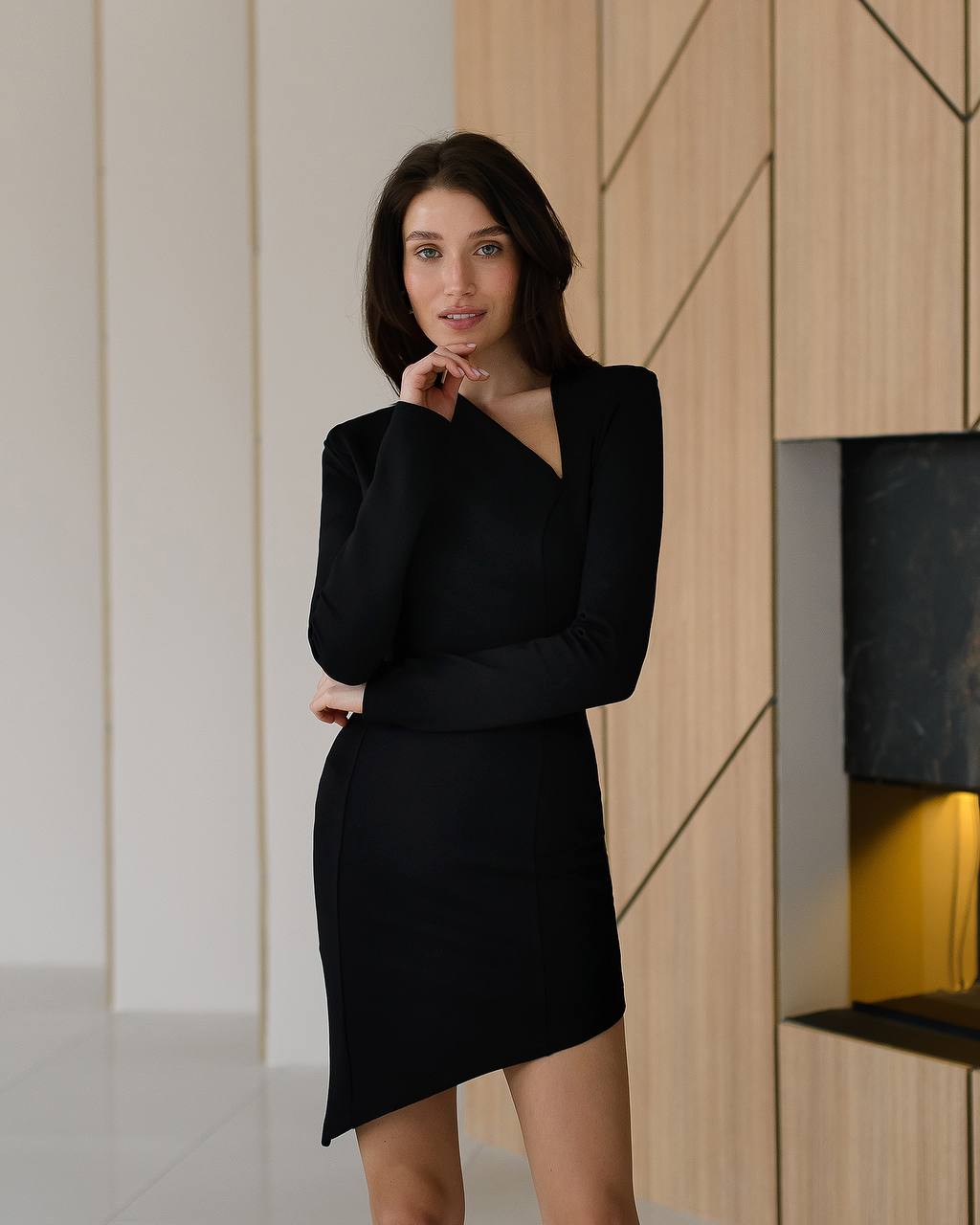 Woman wearing a black dress standing indoors against a modern wall.