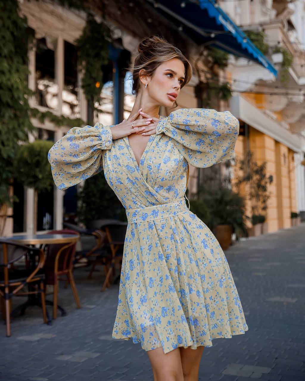 Woman in a floral dress standing on a street with buildings and tables in the background