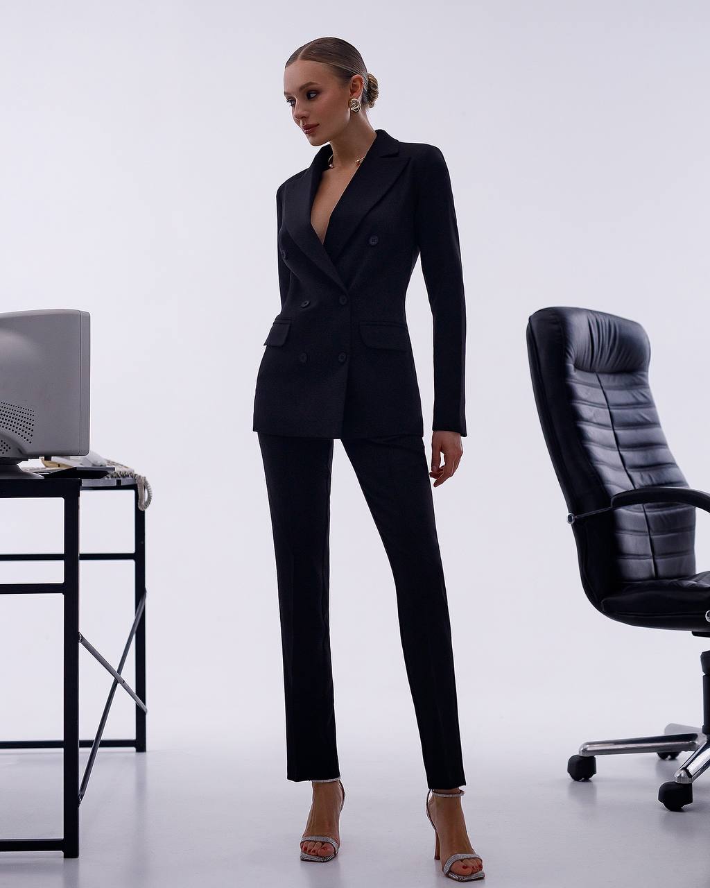 Woman in a black suit standing in an office setting with a chair and desk.