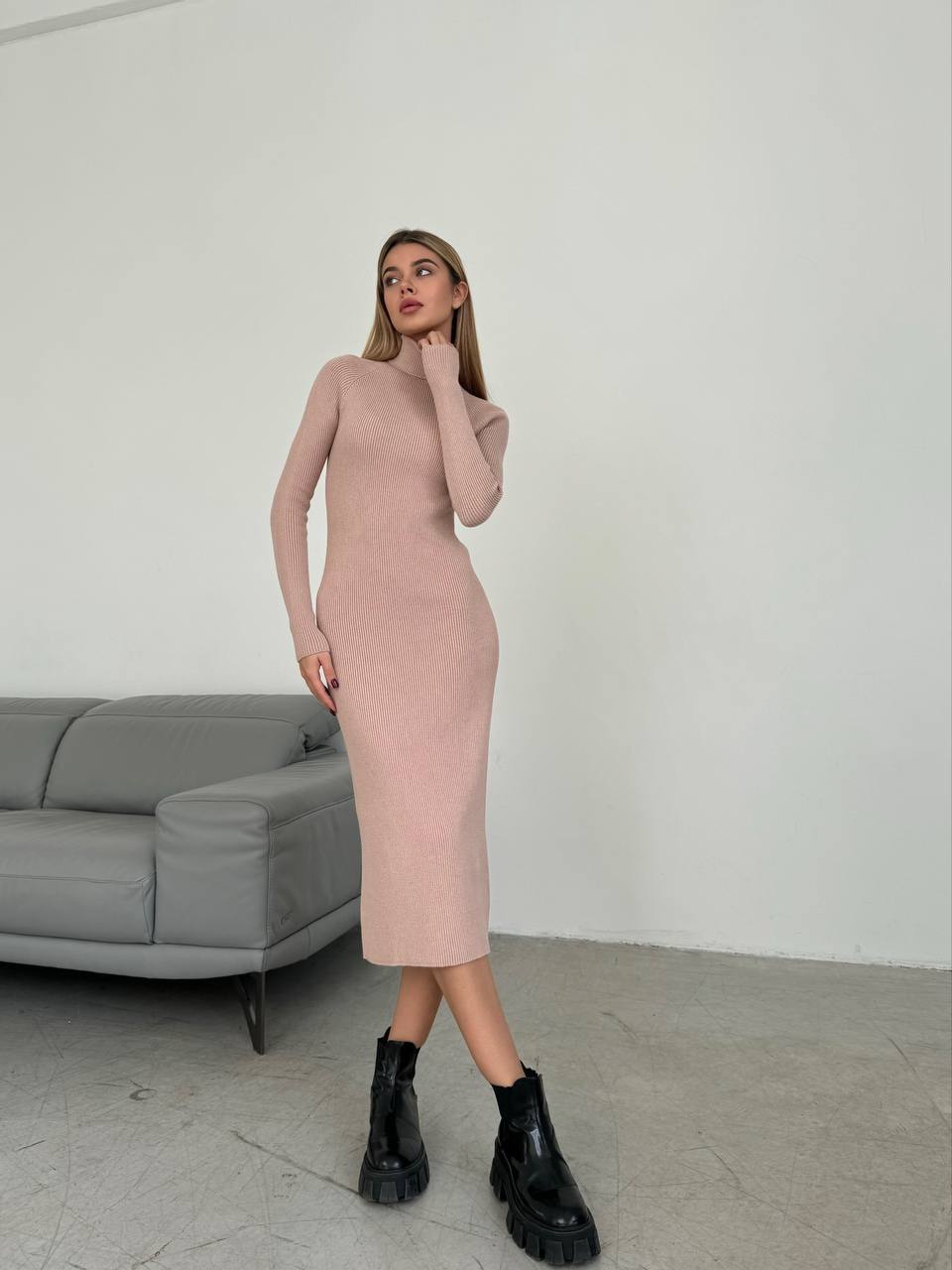 Woman wearing a pink dress and black boots standing in a minimalistic room with a gray sofa.