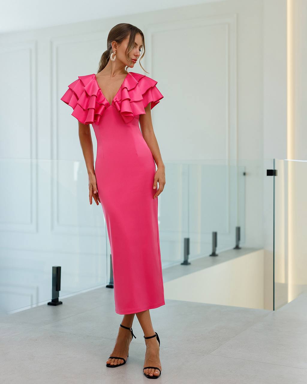 Woman wearing a bright pink dress with ruffled shoulders in a modern indoor setting.