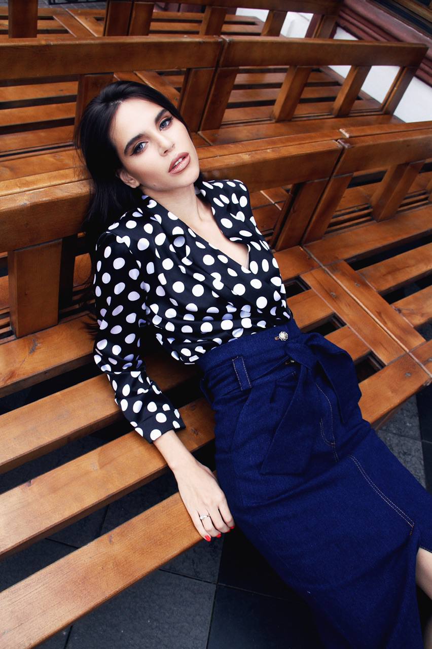 Woman in a polka dot top and blue skirt sitting on a wooden bench.