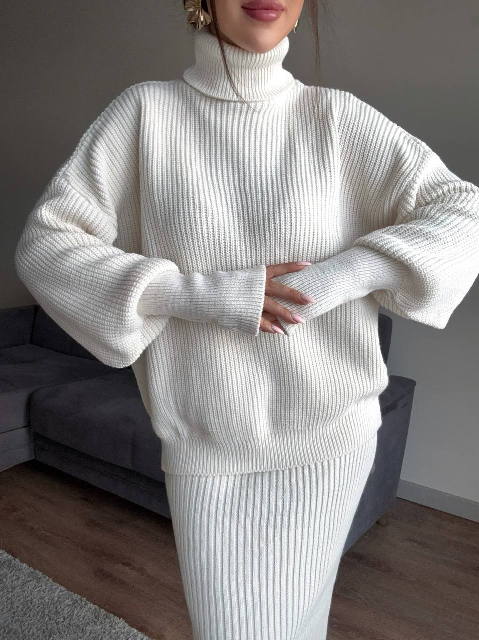 Person wearing a white ribbed sweater and pants in a room with a gray sofa.