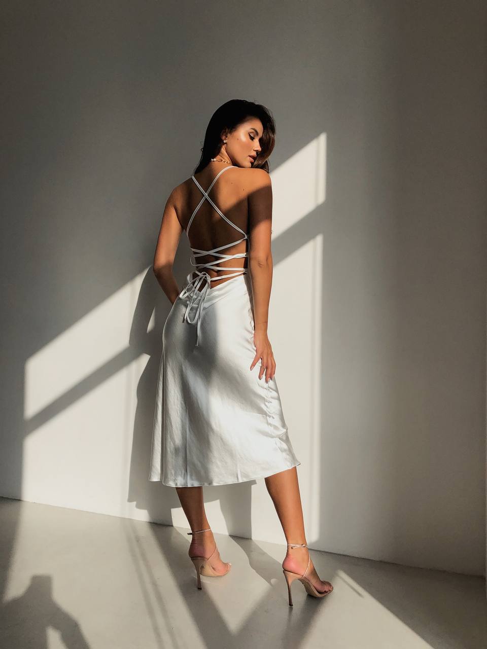 Woman in a white dress standing in a sunlit room with shadows on the wall.
