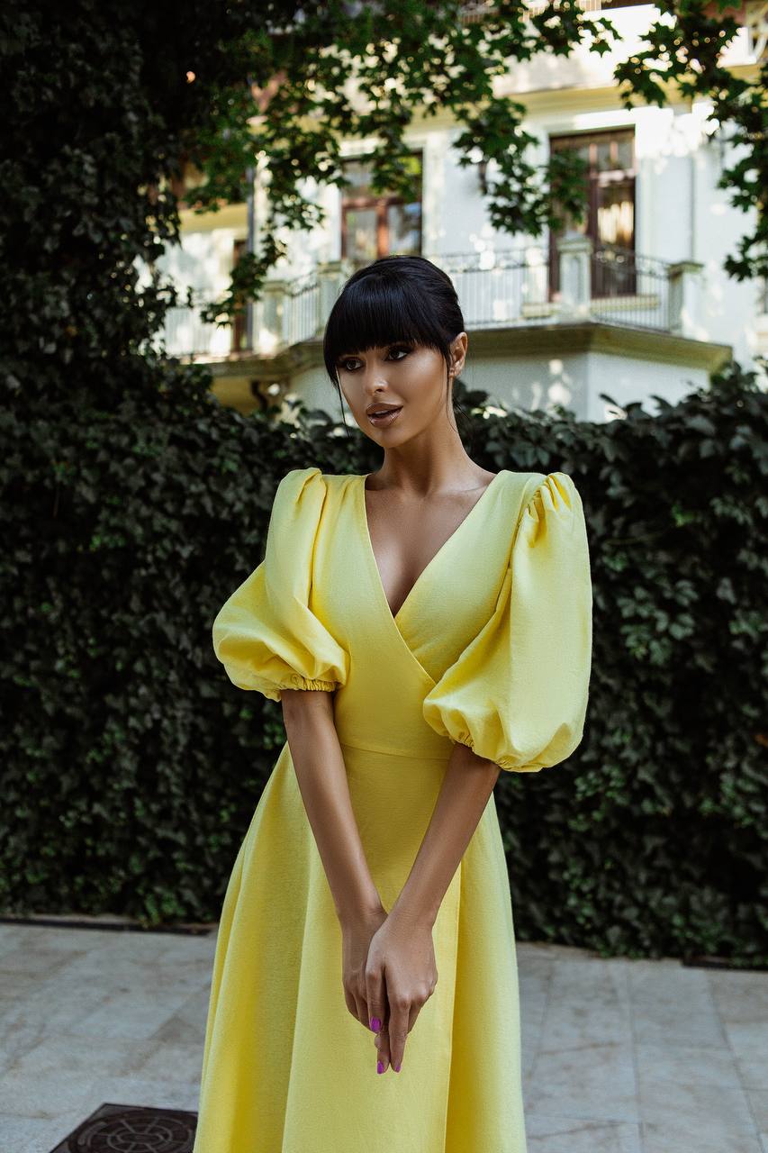 Woman in a yellow dress standing outdoors with greenery and a building in the background