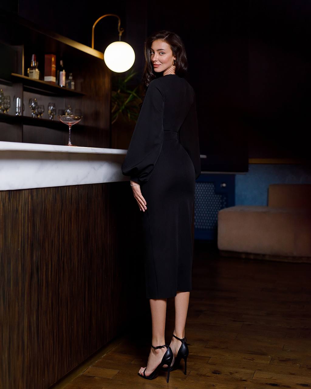 Woman in a black dress standing in a bar setting with dim lighting.